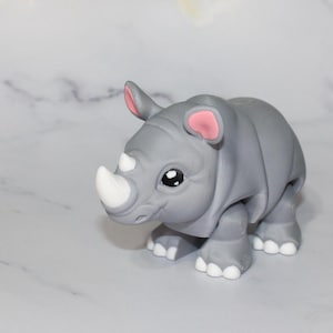 May include: A small gray rhinoceros figurine with a white horn and pink ears. The figurine has a cartoonish style with large eyes and a friendly expression.