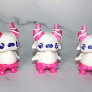 May include: Three small, white figurines with pink and white striped horns, blue eyes, and pink shorts. The figurines have small arms and a rounded body. They are arranged in a row against a light gray background.