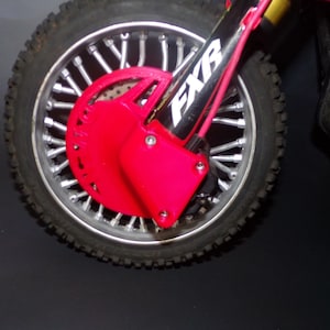 May include: Close-up of a red motorcycle brake disc guard with a silver spoke wheel and black tire. The guard is attached to a black motorcycle fork with the word "FXR" printed on it.