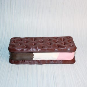 May include: A chocolate ice cream sandwich with a textured, dark brown cookie exterior and layers of brown, white, and pink ice cream. The dessert is presented on a light blue background.