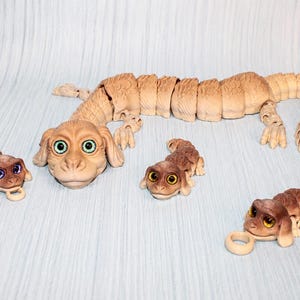 May include: A large articulated beige creature figurine with green eyes and a long segmented body, accompanied by three smaller matching articulated figurines, two of which feature small circular loops attached to their front ends.