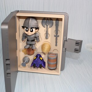 May include: A gray toy set featuring a cartoon knight figure, an axe, a sword, a barrel, and other accessories. The knight is wearing gray armor and a helmet. The set is displayed in an open gray box.
