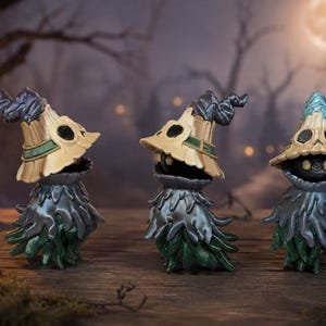 May include: Three small figurines featuring a dark, round body with glowing yellow eyes, wearing a tan, straw-like pointed hat with dark accents and a tattered, metallic grey and green cloak that hangs down to the base in layered, jagged strips.