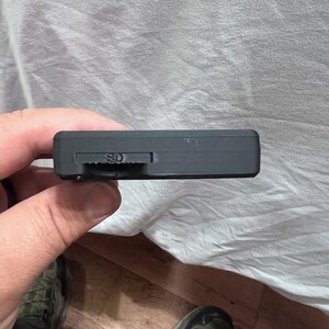 May include: A black rectangular device with an SD card slot is held in a hand. The device has a small slider and the letters "SD" are visible. The background is a wrinkled white sheet and a wooden floor.