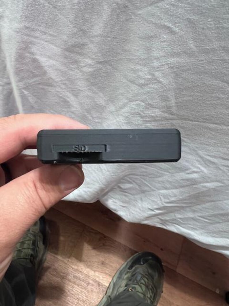 May include: A black rectangular device with an SD card slot is held in a hand. The device has a small slider and the letters "SD" are visible. The background is a wrinkled white sheet and a wooden floor.