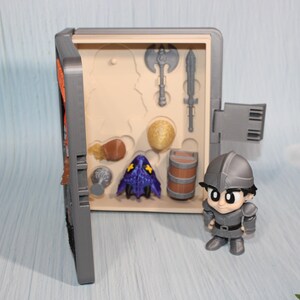 May include: A gray toy chest with a hinged door, open to reveal miniature toy weapons and accessories. A small, gray knight figurine stands beside the chest. The chest contains a miniature axe, sword, and other toy items.