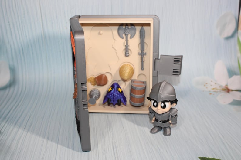 May include: A gray toy chest with a hinged door, open to reveal miniature toy weapons and accessories. A small, gray knight figurine stands beside the chest. The chest contains a miniature axe, sword, and other toy items.