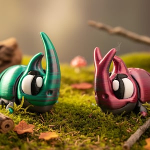 May include: Two 3D-printed beetle figurines, one green and one red, with large cartoon eyes and curved horns. They are set on a bed of green moss with miniature red and white mushrooms and small wooden branches.