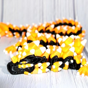May include: A 3D-printed sculpture of a stylized creature, resembling a dragon or dinosaur, made from orange, yellow, black, and white materials. The creature is posed on a white wooden surface, with a focus on its textured, candy corn-like scales.