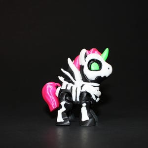 May include: A black and white unicorn figurine with a skeleton design. The unicorn has a pink mane and tail, a green horn, and green eyes. The figurine is set against a black background.