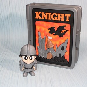 May include: A gray toy knight figure stands next to a gray case with the word "KNIGHT" in orange. The case features a castle and dragon design against an orange background. The knight figure is wearing a helmet and armor.
