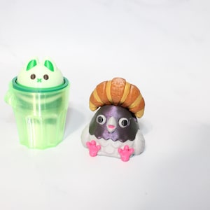May include: A small light green translucent cup containing a pale green cat figure with dark eyes and a small green cross mouth, positioned next to a gray pigeon figure wearing a brown croissant on its head with bright pink feet and beak.