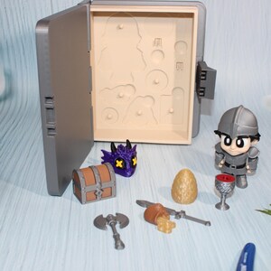 May include: A gray toy box with an open lid, revealing cutouts. Miniature figurines and accessories are arranged around the box, including a knight, dragon head, treasure chest, axe, egg, and goblet. The items are in shades of gray, brown, purple, and gold.