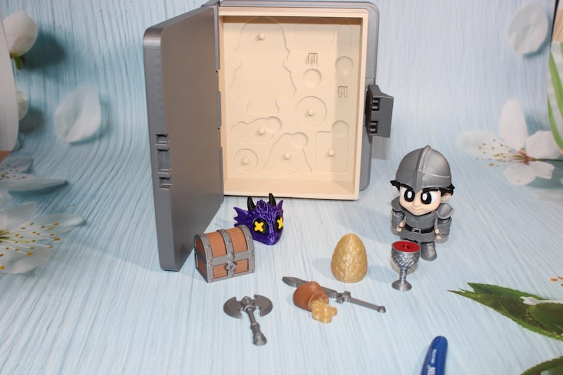 May include: A gray toy box with an open lid, revealing cutouts. Miniature figurines and accessories are arranged around the box, including a knight, dragon head, treasure chest, axe, egg, and goblet. The items are in shades of gray, brown, purple, and gold.