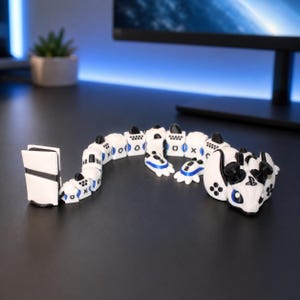 May include: A white and black articulated dragon figurine constructed from gaming controller components. The creature features a head with PlayStation symbols, a segmented body with controller buttons, and a tail shaped like a game console.