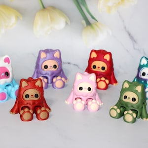 May include: A collection of seven colorful cat-themed figurines. Each figurine is in a seated position and features a cat-like hood and paws. The color palette includes red, pink, green, blue, purple, and white, with contrasting details.