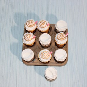 May include: Nine cupcakes in a brown cardboard container, some with white frosting and a caramel swirl, others with white lids. Pink straws are inserted into some of the cupcakes. The background is a light blue surface.