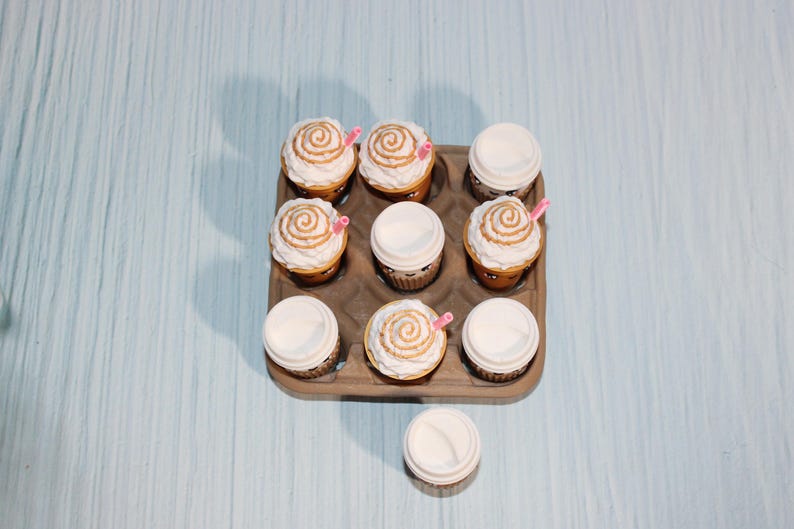 May include: Nine cupcakes in a brown cardboard container, some with white frosting and a caramel swirl, others with white lids. Pink straws are inserted into some of the cupcakes. The background is a light blue surface.