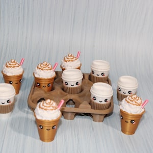 May include: A collection of miniature coffee cups and lidded cups with cute, smiling faces. Some cups have whipped cream and a pink straw. The cups are arranged in a brown cardboard carrier, set against a light blue background.