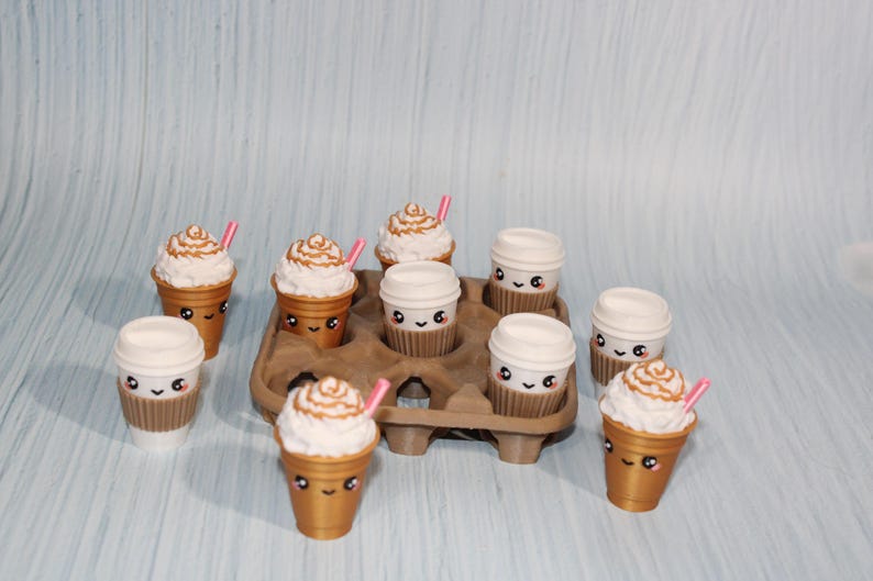 May include: A collection of miniature coffee cups and lidded cups with cute, smiling faces. Some cups have whipped cream and a pink straw. The cups are arranged in a brown cardboard carrier, set against a light blue background.