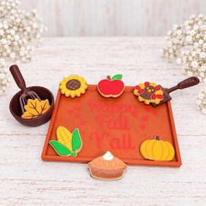 May include: A rectangular orange tray with the text Happy Fall Y'all, featuring autumn-themed cookies shaped like a sunflower, apple, turkey, corn, pumpkin, and pie. A brown bowl contains a leaf-shaped cookie and a brown spatula is included.