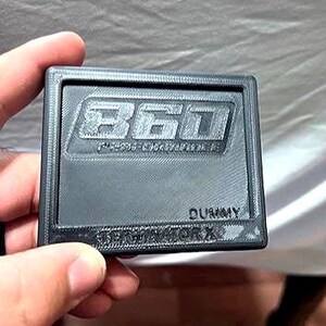 May include: A dark gray rectangular object with the number "360" embossed in the center. The word "DUMMY" is below the number, and "PERFORMANCE WORX" is at the bottom. The object is held by a hand.