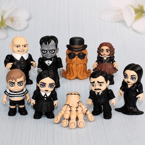 May include: A collection of eight miniature figurines representing characters from a gothic family, including a bald man, a grey-skinned man, a hairy creature with a hat, a girl with brown hair, a boy in a striped shirt, a girl with braids, a hand, and a woman.