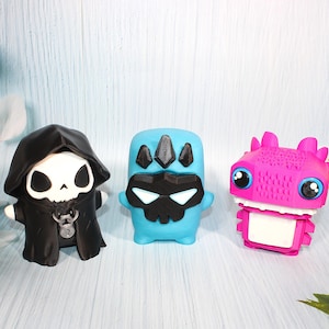 May include: Three small figurines: a black hooded figure with a skull face and a silver padlock pendant, a light blue character with a black skull mask and crown, and a bright pink creature with large eyes and a textured head.