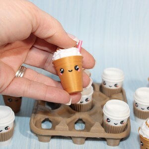 May include: A collection of miniature coffee drinks with cute, smiling faces. The drinks are in brown cups with white whipped cream and pink straws. Some are in a brown egg carton.