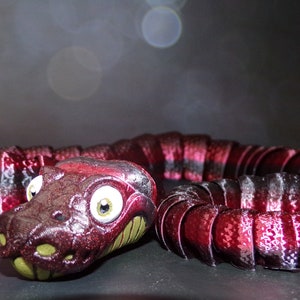 May include: A segmented, red and black toy snake with a detailed head and large, round eyes. The snake's body is made of connected segments, and the head has a detailed, textured appearance. The toy is likely a decorative item or a child's plaything.