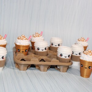 May include: A collection of adorable coffee cup figurines, some with whipped cream and pink straws. The cups are white and gold, with cute faces and brown sleeves. They are arranged in a brown cardboard carrier.