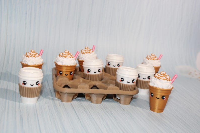 May include: A collection of adorable coffee cup figurines, some with whipped cream and pink straws. The cups are white and gold, with cute faces and brown sleeves. They are arranged in a brown cardboard carrier.