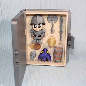 May include: A gray toy box with an open door revealing a knight figurine and miniature accessories. The accessories include a battle axe, sword, barrel, and a purple hooded figure. The knight is wearing gray armor.