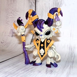 May include: A small, white, goat-like figurine with purple horns and hooves. It is wearing a black and gold robe with a skull design and holding a purple and gold staff with a skull at the top.