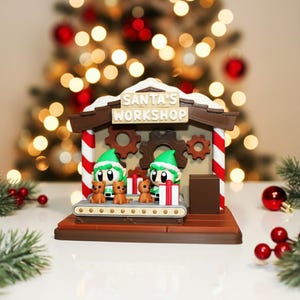 May include: A Christmas-themed figurine of Santa's Workshop. The figurine features two elves in green hats, teddy bears, and a conveyor belt. The workshop has a brown roof with the text "SANTA'S WORKSHOP". Red and white striped poles frame the scene.