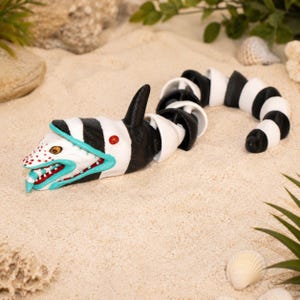May include: A segmented, articulated creature figure featuring a black and white striped body, a white head with red spots, a teal-colored mouth interior with white teeth, and a single dorsal fin. The figure has a long, flexible, snake-like tail.