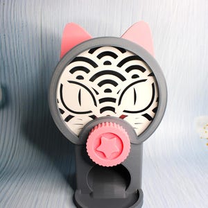 May include: A gray and pink cat-shaped phone stand. The stand features a circular design with cat eyes and a wave pattern, pink ears, and a pink star-shaped knob. The stand is designed to hold a phone.