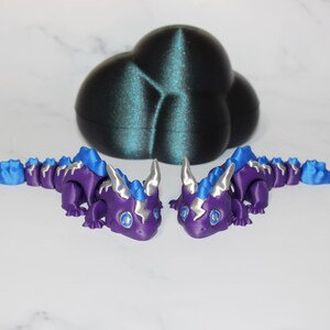 May include: Two purple and blue articulated dragon figurines with silver horns and accents are displayed in front of a dark teal cloud-shaped object. The dragons have blue eyes and are posed facing each other.
