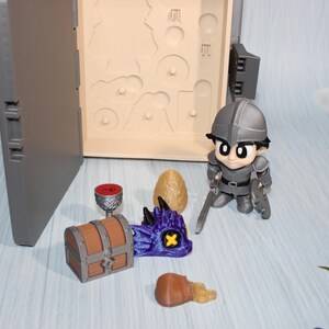 May include: A miniature fantasy playset featuring a knight figurine, a treasure chest, a dragon head, a goblet, an egg, and a treasure chest. The set includes a gray case with a compartment for the figurines. The knight is holding two swords.