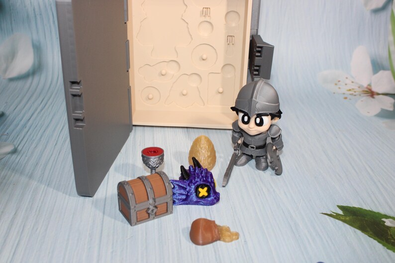 May include: A miniature fantasy playset featuring a knight figurine, a treasure chest, a dragon head, a goblet, an egg, and a treasure chest. The set includes a gray case with a compartment for the figurines. The knight is holding two swords.