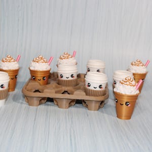 May include: An assortment of miniature coffee cups and iced coffee drinks with whipped cream and pink straws. The cups have cute, smiling faces. Some are in a brown egg carton.