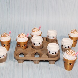 May include: A collection of miniature coffee cups and iced coffees with cute faces. Some have whipped cream and pink straws. They are arranged in a brown egg carton on a light blue surface.