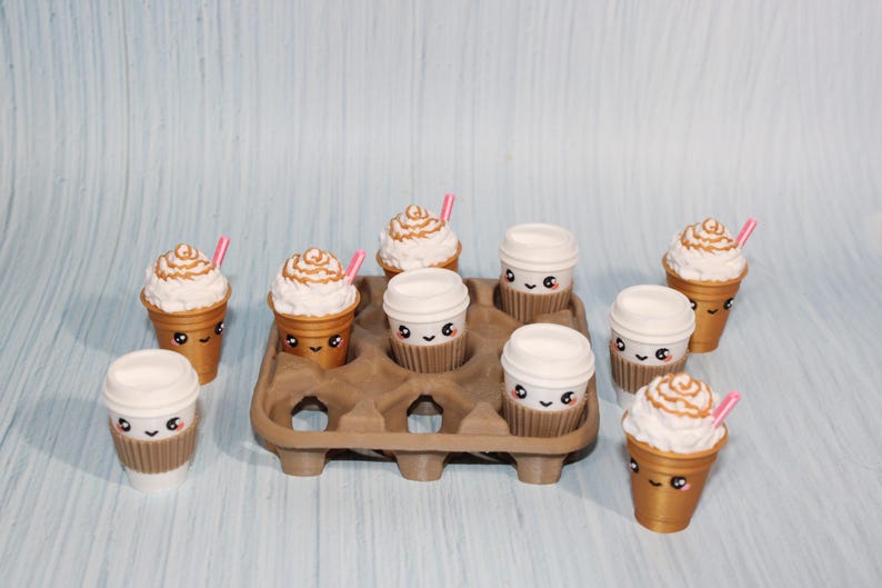 May include: A collection of miniature coffee cups and iced coffees with cute faces. Some have whipped cream and pink straws. They are arranged in a brown egg carton on a light blue surface.