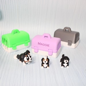 May include: Three miniature pet carriers in bright green, light purple, and dark gray, each with a white base and a top handle. The purple carrier features the name MAGGIE printed on the front. Three small black, white, and brown dog figurines.