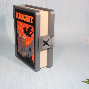 May include: A decorative book with the word "KNIGHT" in orange lettering. The cover features a castle and dragon design against an orange background. The book has a gray frame and a clasp with a sword and crossbones.