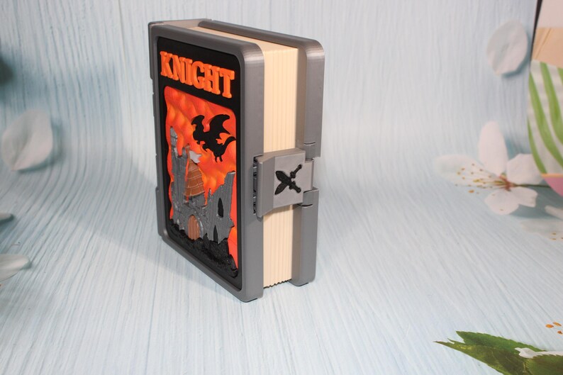 May include: A decorative book with the word "KNIGHT" in orange lettering. The cover features a castle and dragon design against an orange background. The book has a gray frame and a clasp with a sword and crossbones.