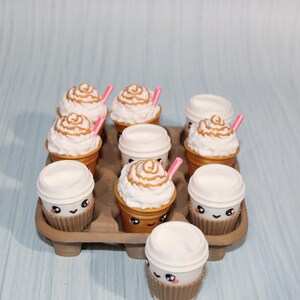 May include: A tray of miniature coffee cups and whipped cream treats. Some cups have lids, others are topped with swirls of cream and caramel drizzle, and pink straws. The cups have cute smiling faces. The tray is brown.