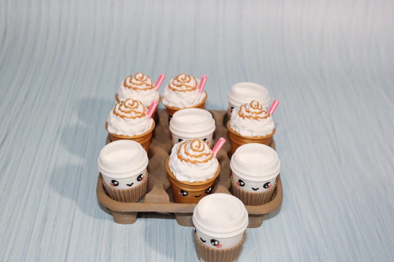 May include: A tray of miniature coffee cups and whipped cream treats. Some cups have lids, others are topped with swirls of cream and caramel drizzle, and pink straws. The cups have cute smiling faces. The tray is brown.