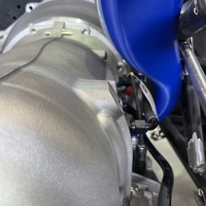May include: Close-up of a silver engine with a blue funnel attached to a black hose. The funnel is used to pour fluids into the engine.