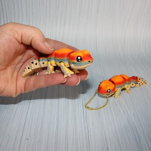 May include: Two articulated lizard figurines featuring a vibrant gradient of orange, yellow, and blue stripes on their bodies. The tails are cream-colored with black spots. One lizard is held in a hand, while the other lies flat with a gold chain.